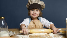 Happy cute little girl flattening pastry using rolling pin while